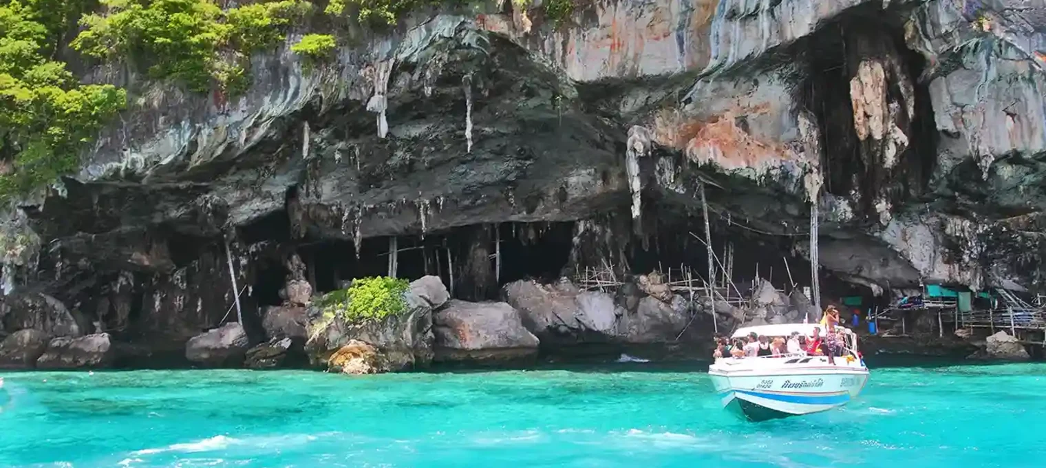 Boat approaching rocky cave entrance near lush green cliffs in Phuket's turquoise waters.