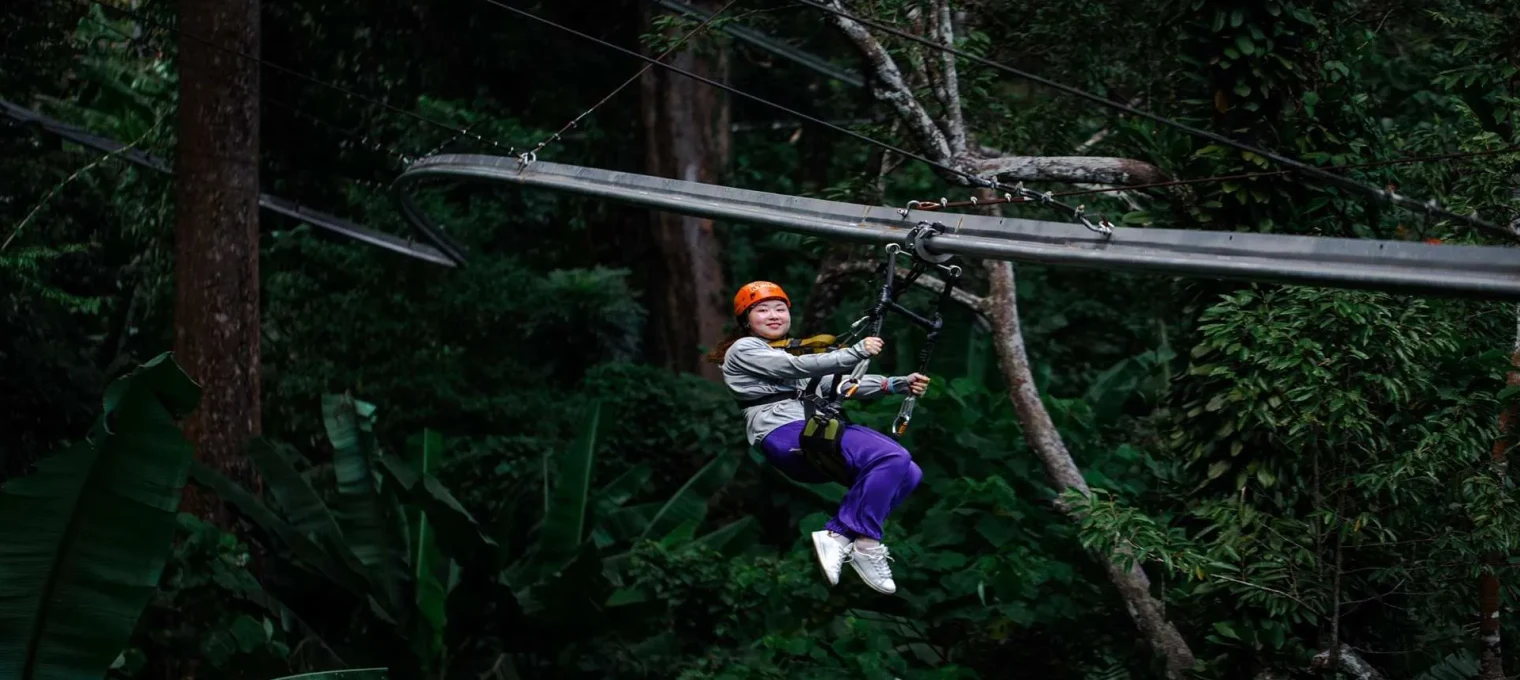 Person ziplining through lush greenery in Phuket, wearing an orange helmet and casual attire.