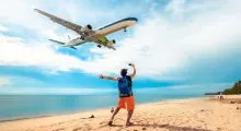 A traveler on a beach in Phuket waves at a plane flying overhead.