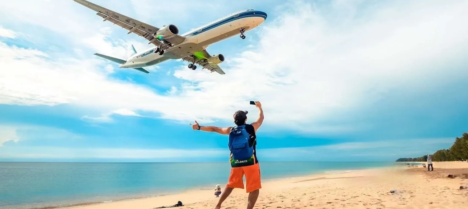 A traveler on a beach in Phuket waves at a plane flying overhead.