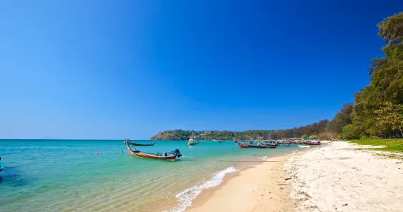 Пляж в Пхукете с прозрачной водой и рыбацкими лодками на фоне яркого неба.