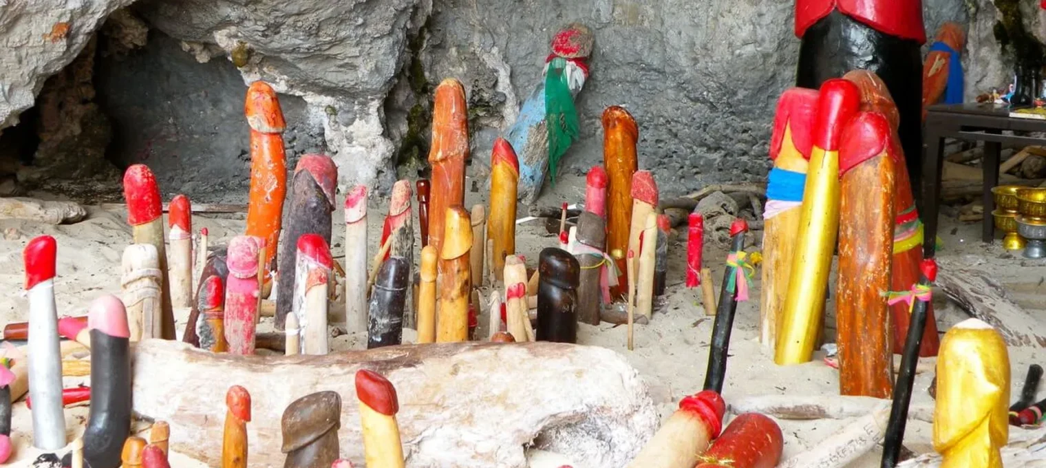 Colorful wooden phallic sculptures scattered on sandy ground in a cave in Phuket.