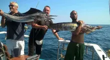 Three men proudly holding large fish on a boat in Phuket's clear blue waters.