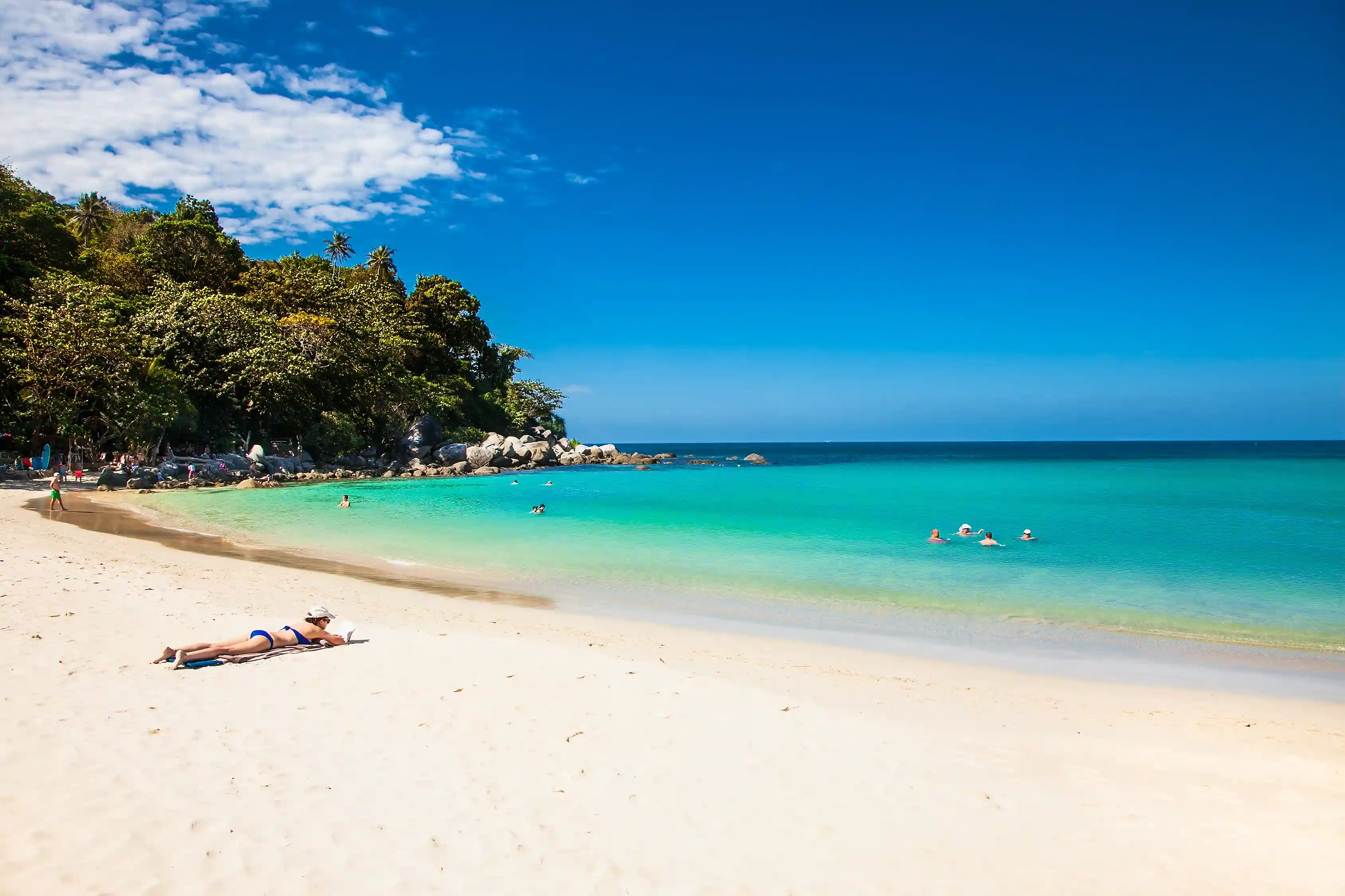 Sunny beach in Phuket with clear blue waters and people swimming and lounging on the sand.