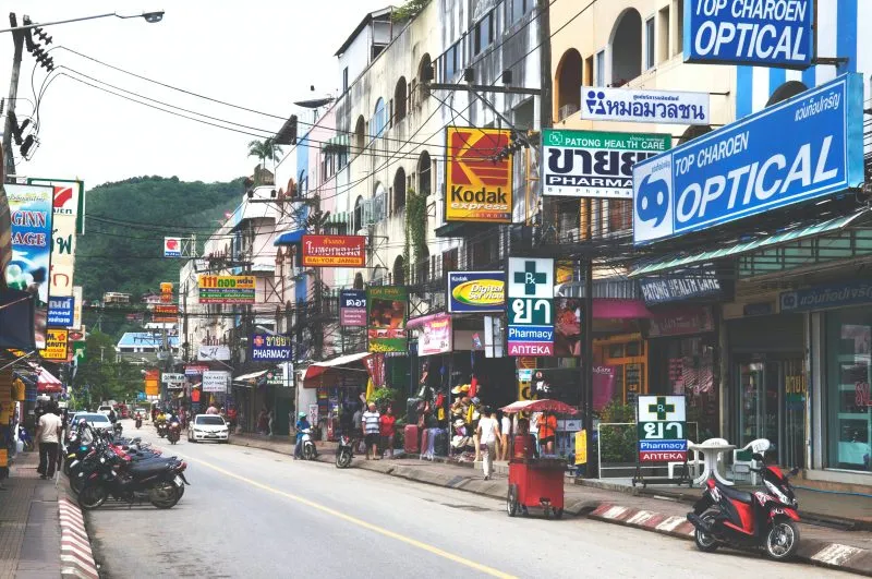 Busy street in Phuket featuring shops, pharmacies, and motorcycles parked alongside.