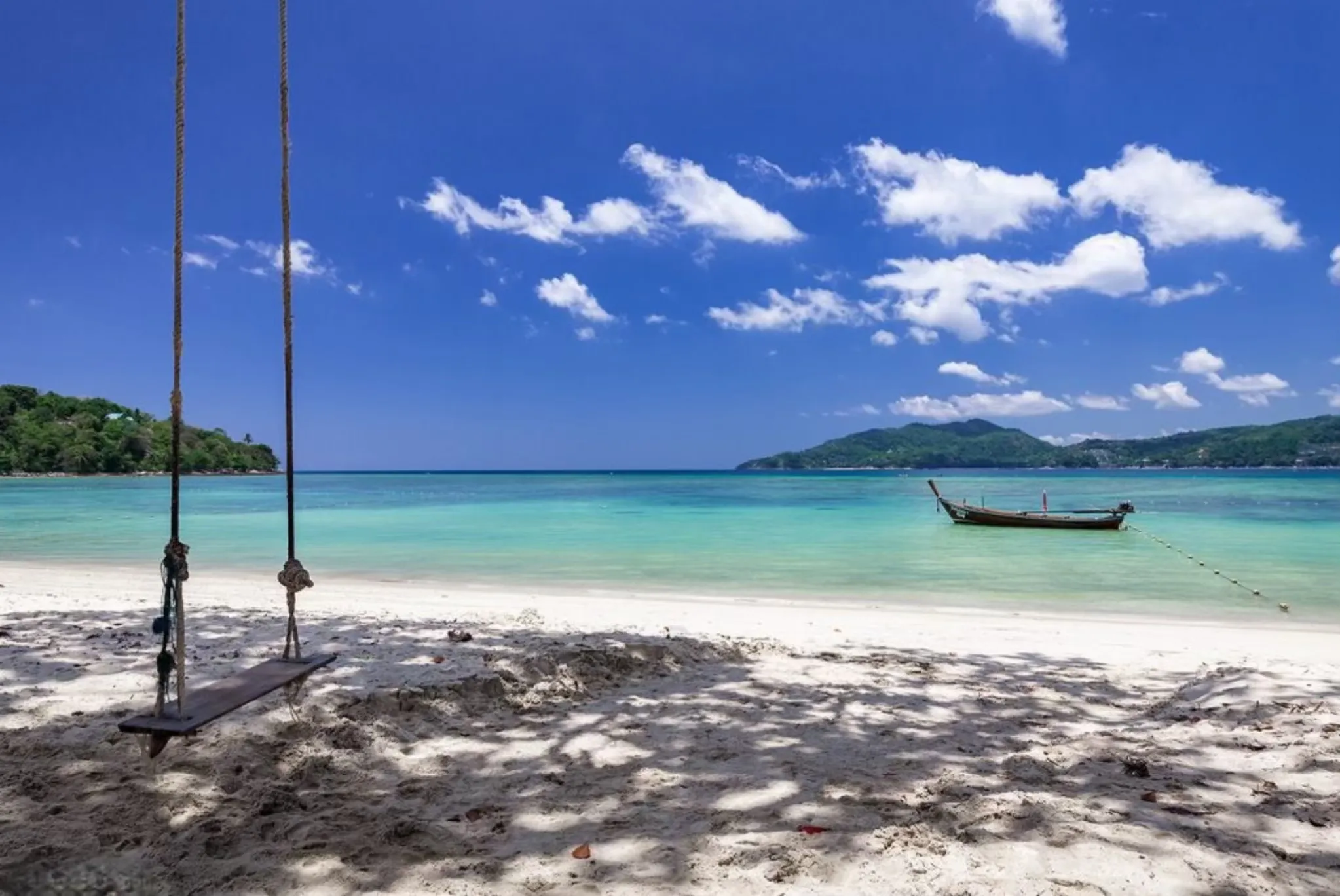A tranquil beach in Phuket with a swing, turquoise water, and a fishing boat in the background.