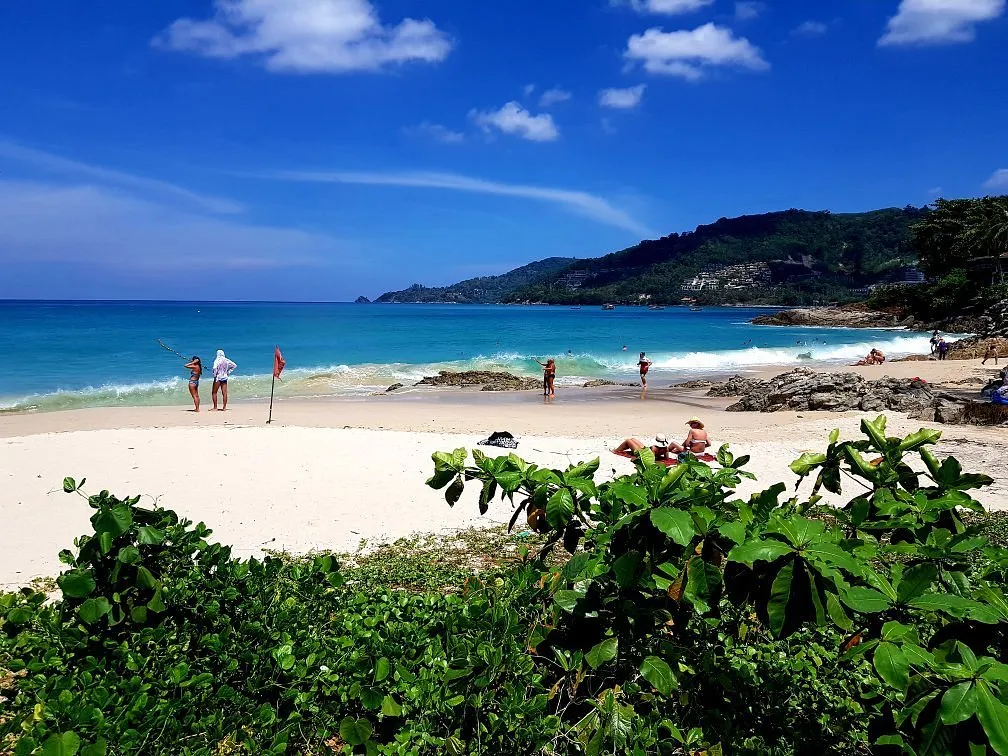 Bright beach in Phuket with people enjoying the sun, gentle waves, and lush greenery.