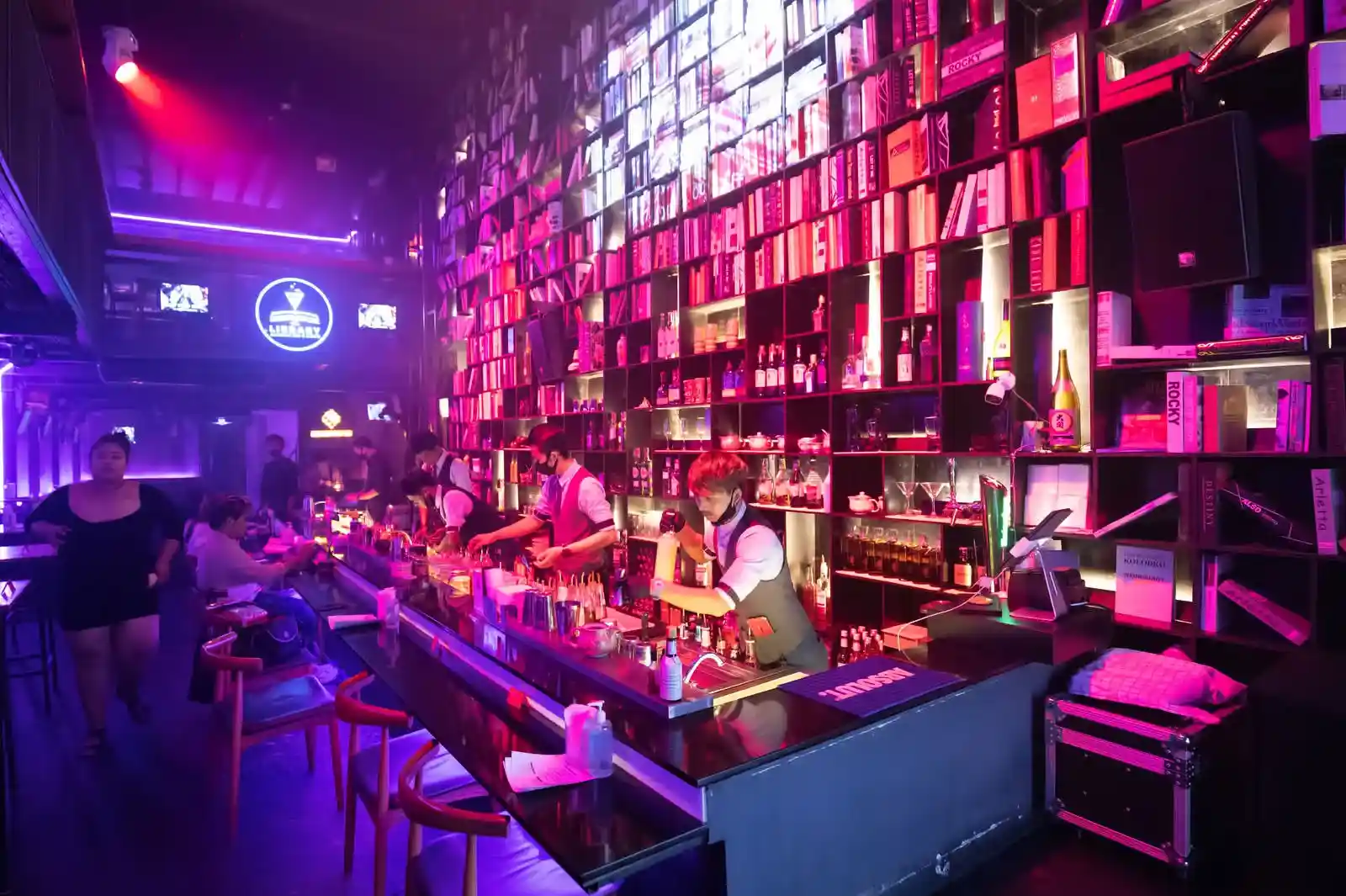 A vibrant bar in Phuket with colorful lights, bookshelves, and bartenders serving drinks.