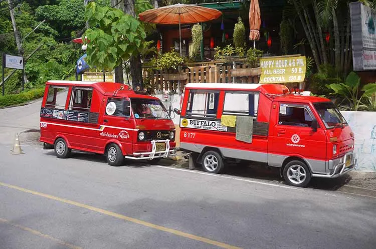 Red tuk-tuks parked on a street in Phuket, Thailand, near a bar and trees.