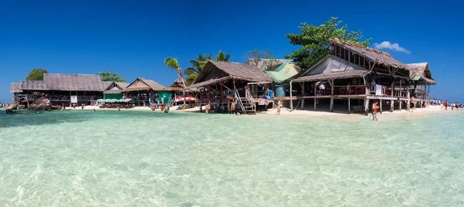 Scenic beachfront view of wooden huts in Phuket, clear turquoise water, and blue skies.