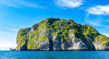 A lush green island rises from clear blue waters near Phuket, Thailand, under a bright sky.