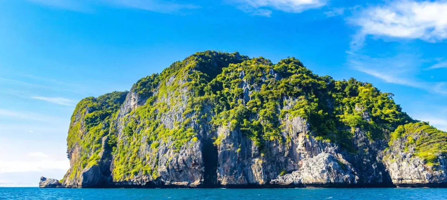 A lush green island rises from clear blue waters near Phuket, Thailand, under a bright sky.
