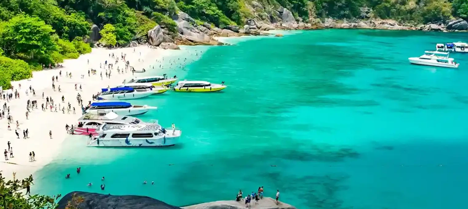 Пляж на Пхукете с яркой водой и множеством катеров на фоне зелёных холмов.