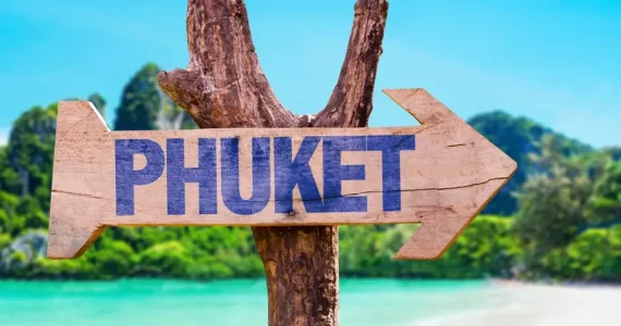 Деревянный указатель с надписью "PHUKET" на фоне пляжа и palm деревьев.