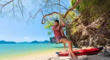 A person swings from a tree by the beach in Phuket, surrounded by turquoise water and hills.