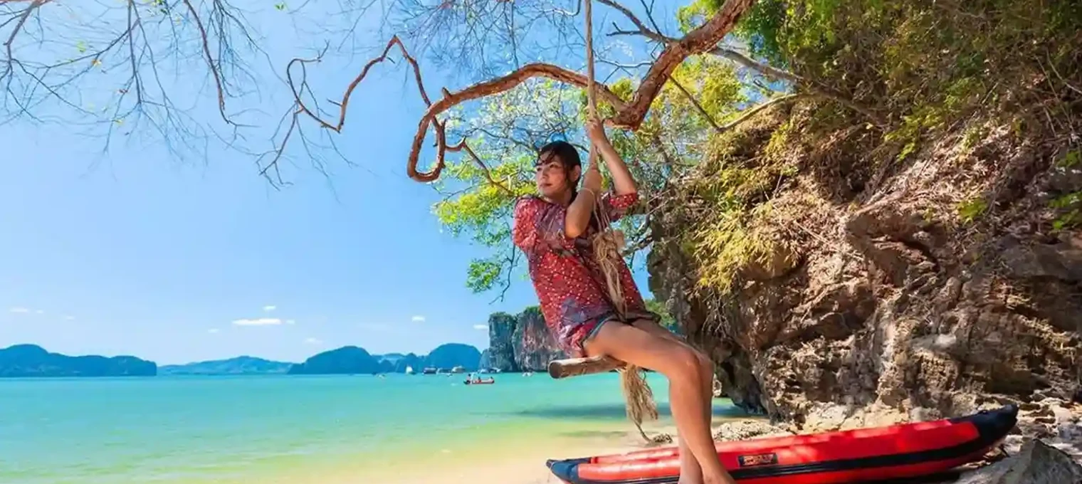 A person swings from a tree by the beach in Phuket, surrounded by turquoise water and hills.
