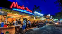 Busy street scene at night outside Mook Dee restaurant in Phuket, featuring illuminated signage.