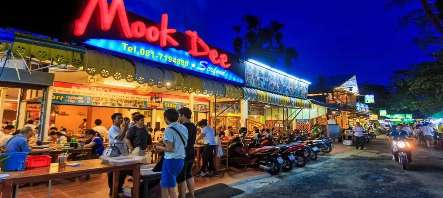 Busy street scene at night outside Mook Dee restaurant in Phuket, featuring illuminated signage.