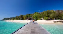 A wooden pier leads to a sandy beach and blue waters in Phuket, surrounded by lush greenery.