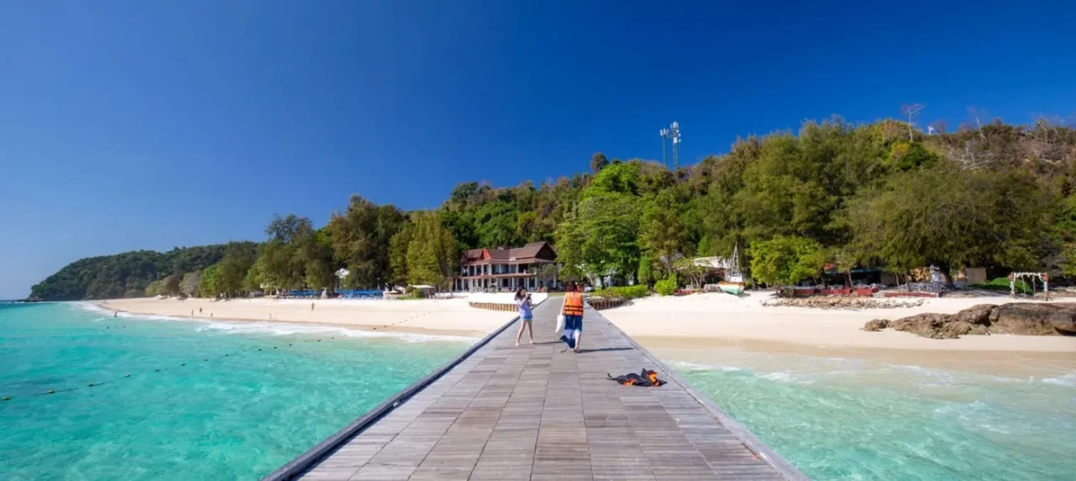 A wooden pier leads to a sandy beach and blue waters in Phuket, surrounded by lush greenery.