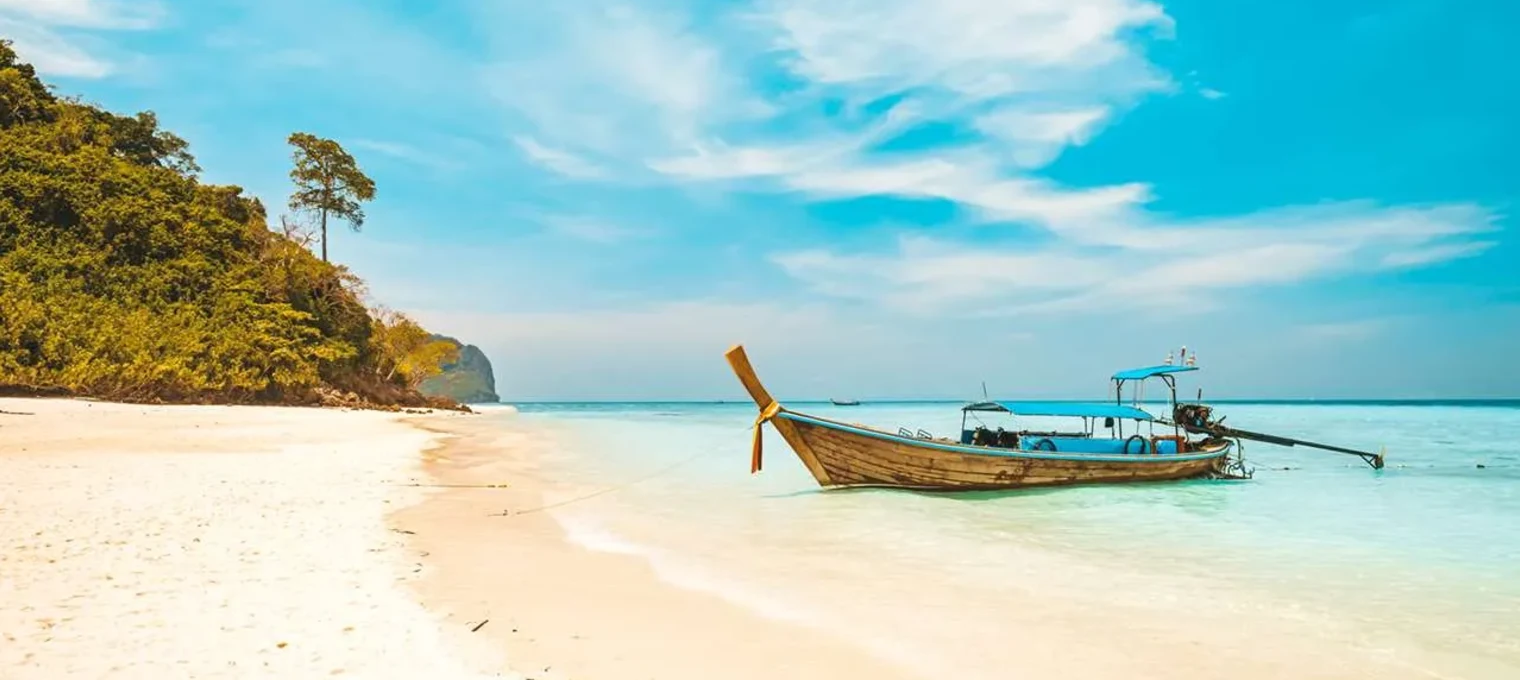 A long-tail boat anchored on a sandy beach in Phuket, surrounded by turquoise waters.