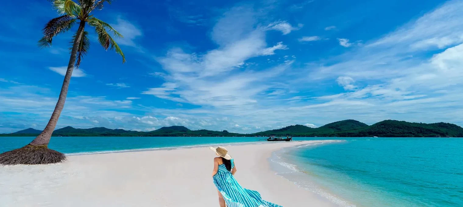 Woman in a blue striped dress walking on a white sandy beach in Phuket, Thailand.