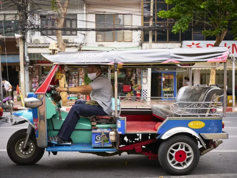 A tuk-tuk driver operating in Phuket, surrounded by local shops and greenery.