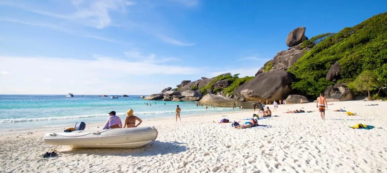 Пляж на Пхукете с белым песком, скалами и отдыхающими под ясным небом.