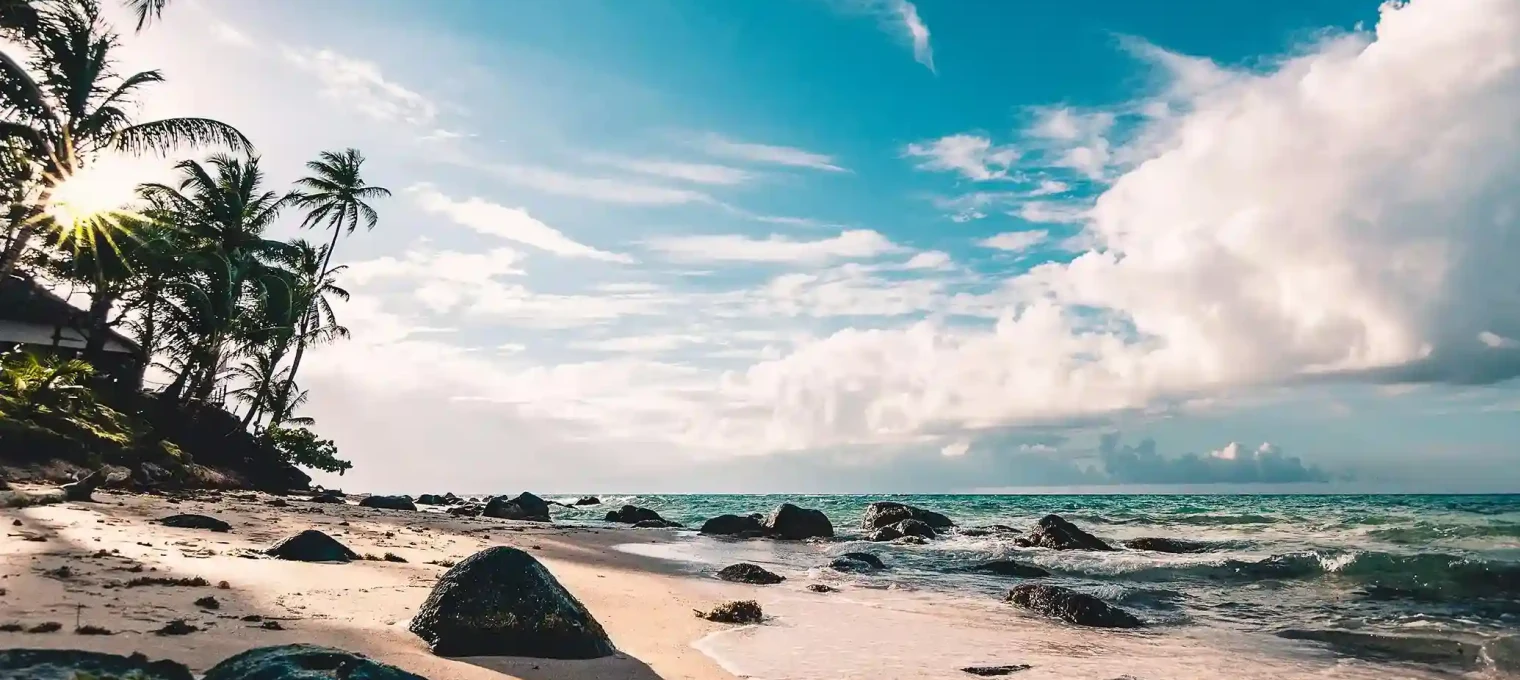 Пляж на Пхукете с камнями, пальмами и небом в облаках. Тёплые волны омывают берег.
