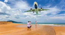 Tourist taking a photo on the beach as a plane flies overhead in Phuket, Thailand.