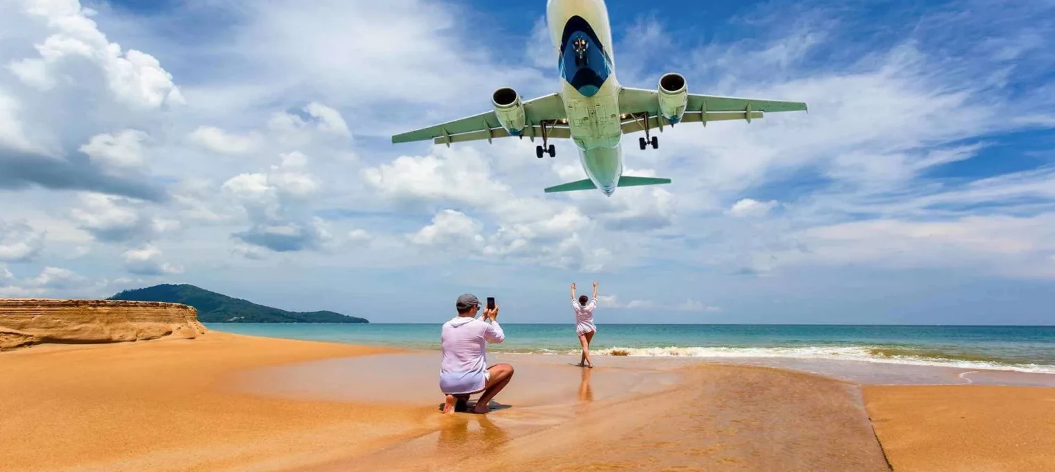 Tourist taking a photo on the beach as a plane flies overhead in Phuket, Thailand.