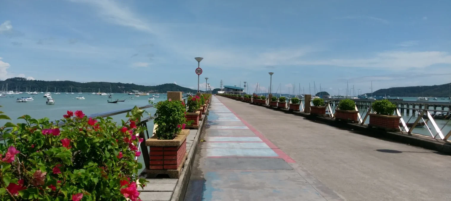 Scenic walkway in Phuket lined with flowers and boats in the harbor.