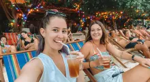 Two smiling women enjoy cocktails on a beach chair in Phuket, surrounded by colorful lights.
