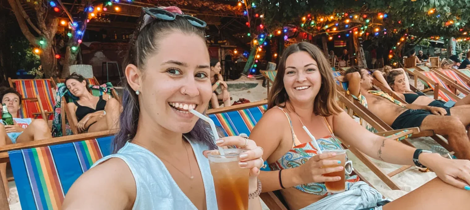 Two smiling women enjoy cocktails on a beach chair in Phuket, surrounded by colorful lights.
