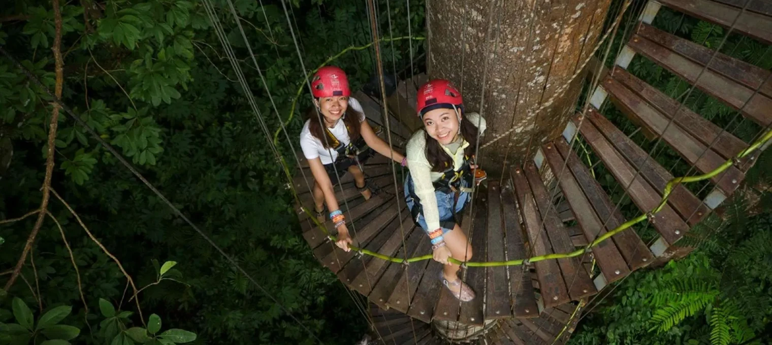 Two women in helmets enjoying a treetop adventure in Phuket, surrounded by lush greenery.