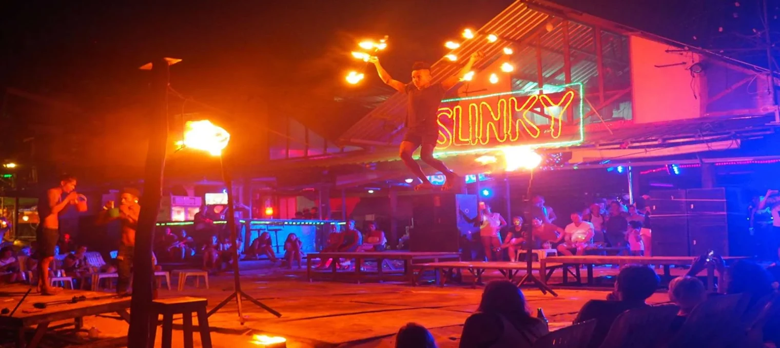 A fire performer leaps with flaming torches at a lively beach bar in Phuket.