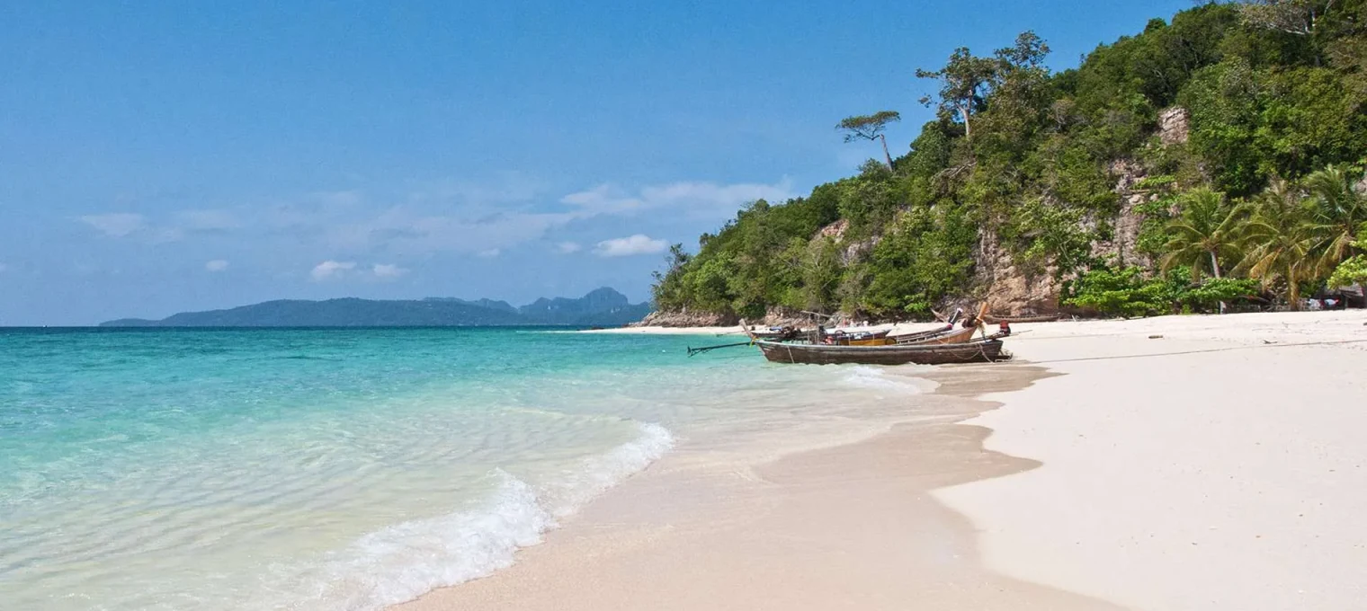 Serene beach in Phuket with clear waters, gentle waves, and fishing boats along the shore.