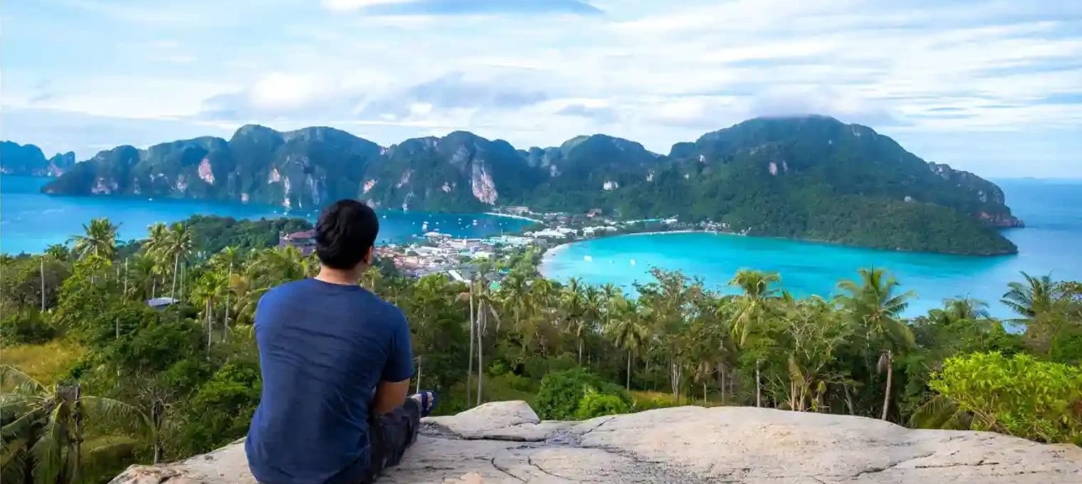 A person sitting on a rock overlooks the stunning coastline of Phuket with lush greenery.