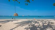 A sunny beach in Phuket with people walking by and boats in the clear water.