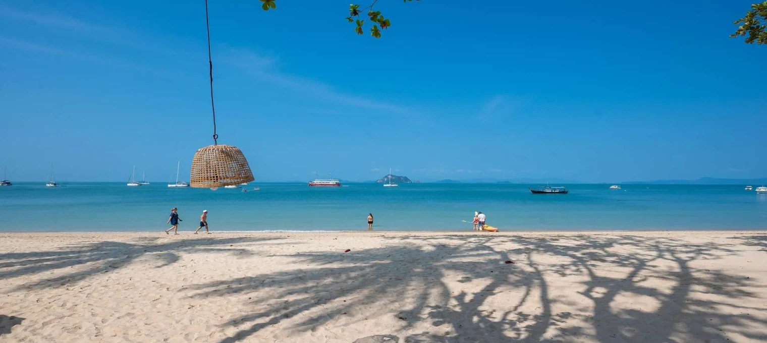 A sunny beach in Phuket with people walking by and boats in the clear water.