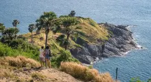 Two people stand on a hill in Phuket, overlooking a rocky coastline and palm trees by the sea.