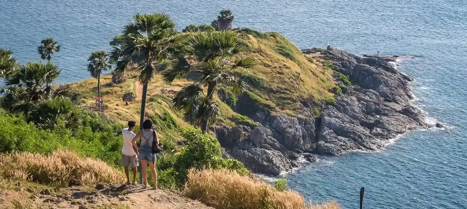 Two people stand on a hill in Phuket, overlooking a rocky coastline and palm trees by the sea.