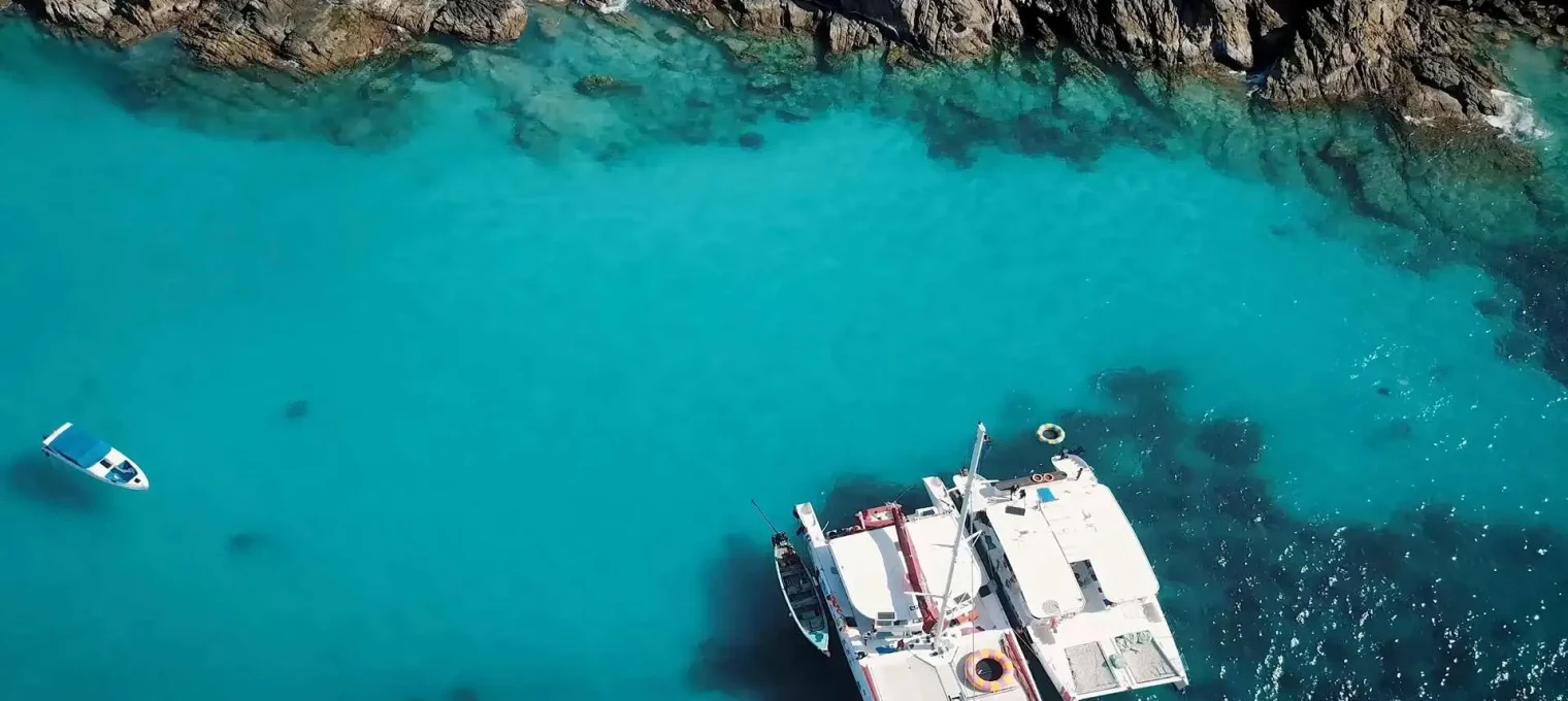 Aerial view of a boat anchored in crystal-clear turquoise waters near rocky coastline in Phuket.