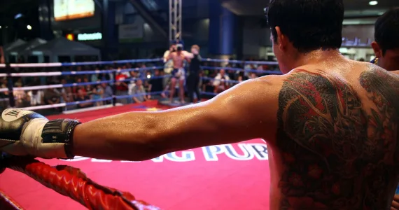A tattooed male fighter rests on the ropes in a vibrant Muay Thai boxing match in Phuket.