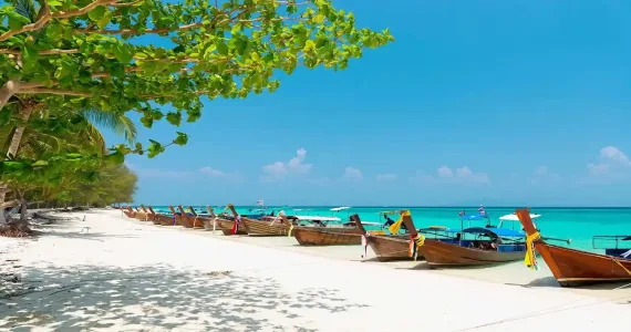 Long-tail boats lined up on a sandy beach in Phuket under a clear blue sky.