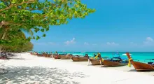 Long-tail boats lined up on a sandy beach in Phuket under a clear blue sky.