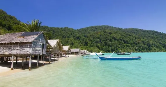 Пейзаж на пляже Пхукета с тропическими домиками и лодками на фоне зелёных холмов.