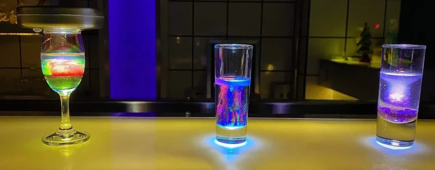 Three illuminated glasses on a bar counter in Phuket, showcasing colorful drinks.