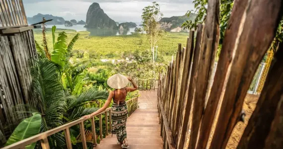 Woman in traditional hat walking down wooden stairs in lush Phuket landscape.
