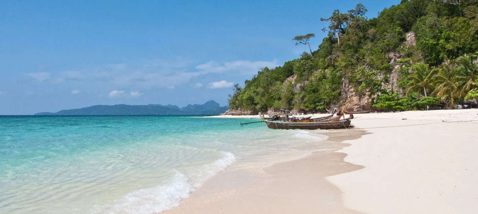 Serene beach in Phuket with clear water, sandy shore, and traditional boats along the coast.